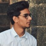 A stylish young man in glasses leans against a brick wall wearing a casual light blue shirt.